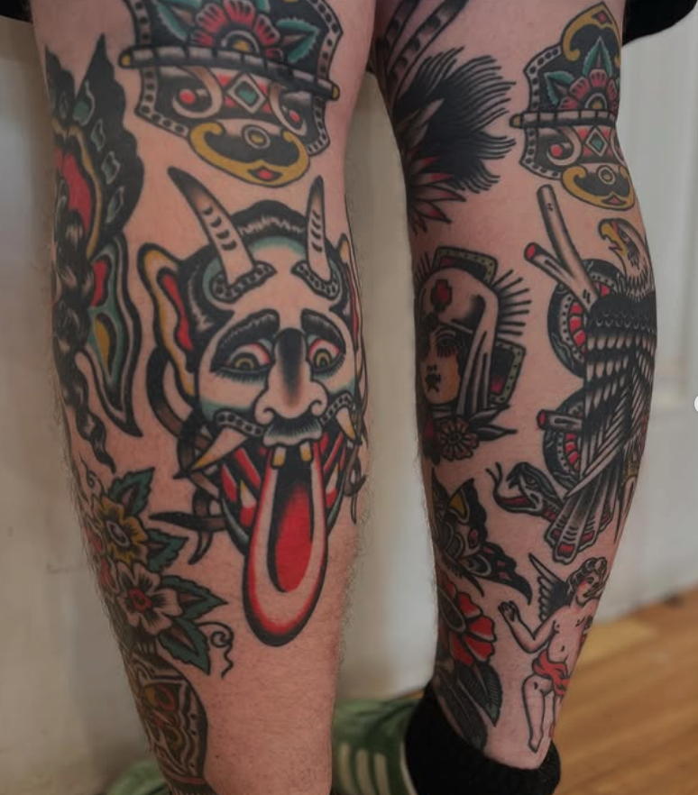 Colorful tattoos on person's legs and arms, including a demon-like face with horns, a woman's face in a folk art style, a bird, and floral patterns.