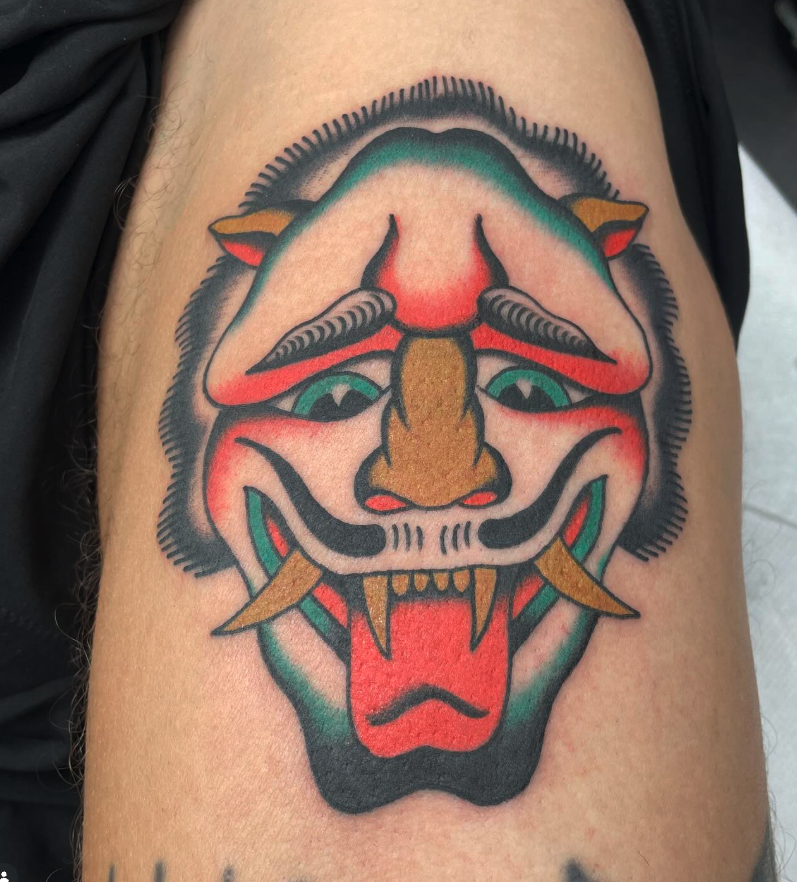 Tattoo of a traditional Japanese mask with a fierce expression, sharp teeth, and colorful details in black, red, and yellow.