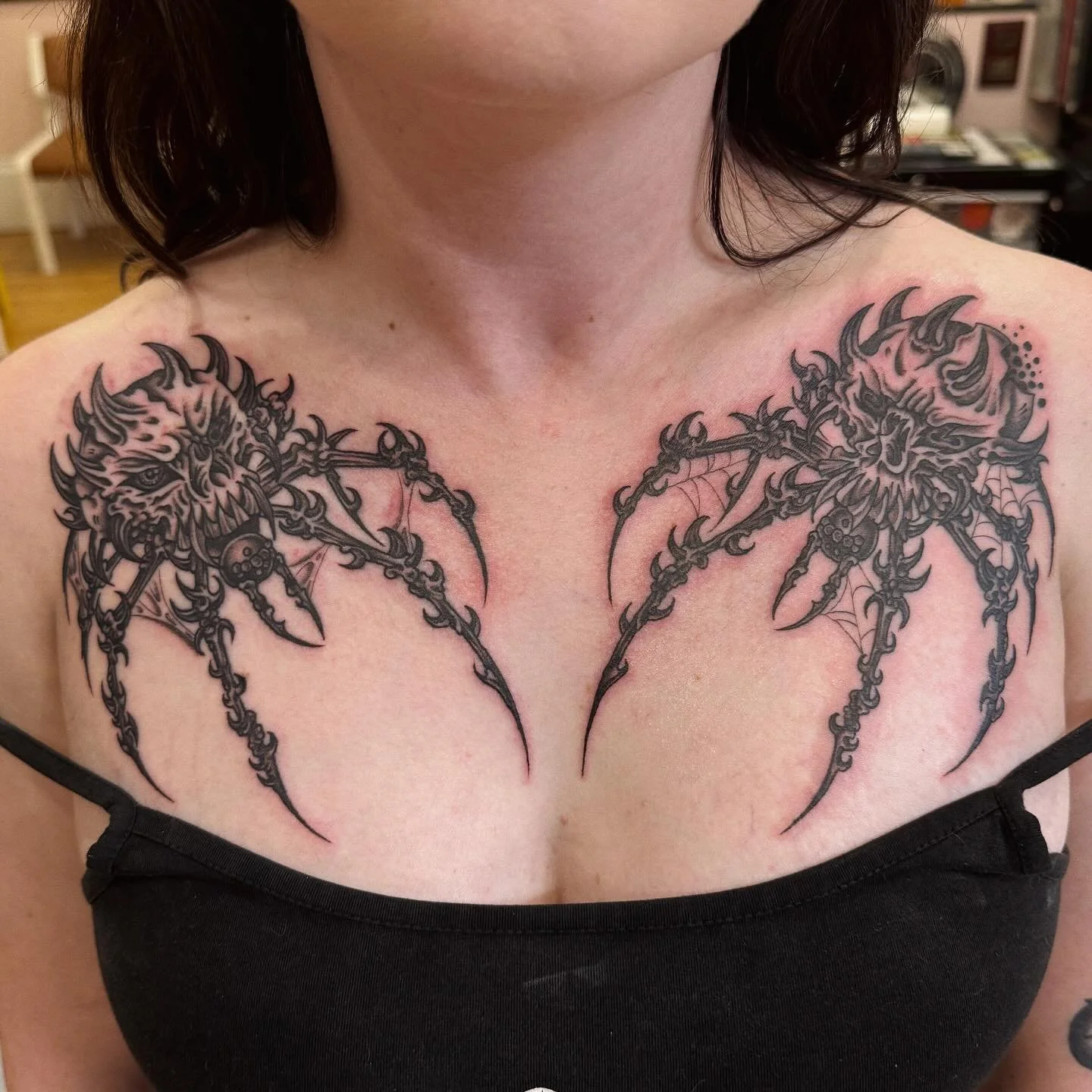 Tattoo of two demonic faces with sharp features, horns, and fangs, connected by web-like structures and claws, covering the upper chest and shoulders.