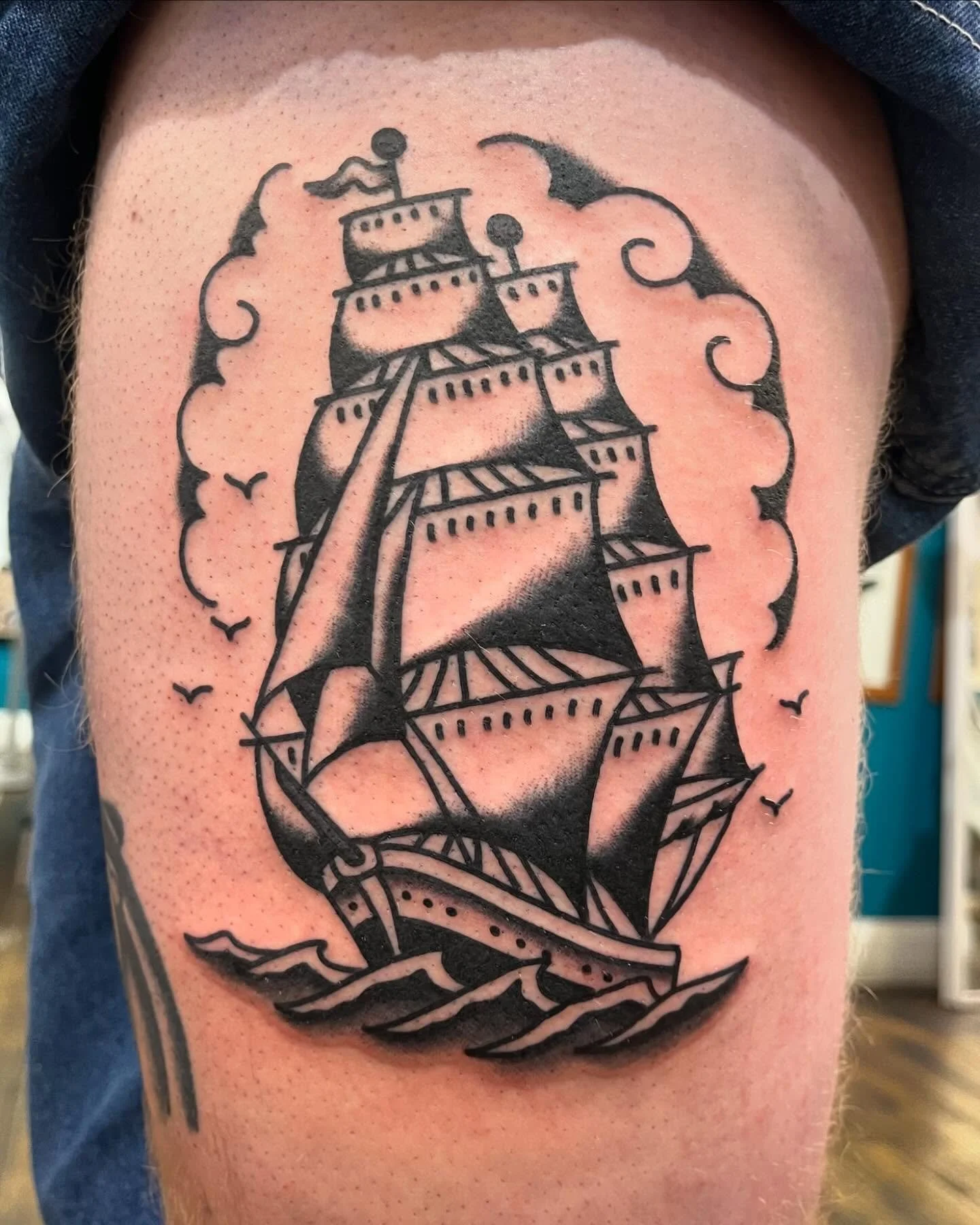 Black outline tattoo of a sailing ship on the water, surrounded by clouds and flying birds.
