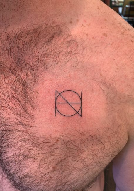A small black tattoo of a geometric design on a person's upper chest near the shoulder, with chest hair nearby.