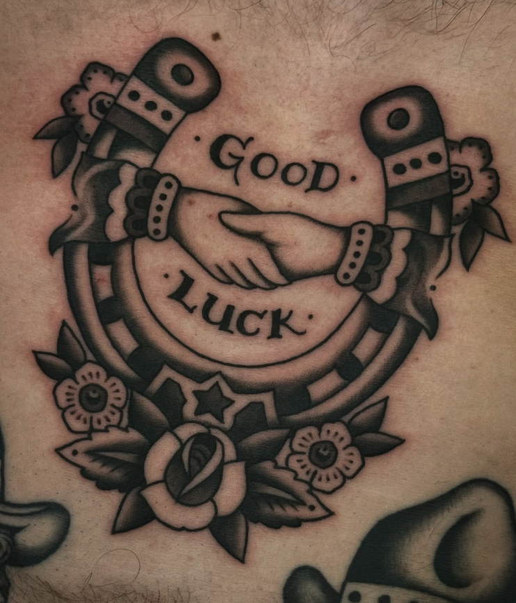 Traditional tattoo featuring two clasped hands, surrounded by flowers, with the words "Good Luck" written above and below.