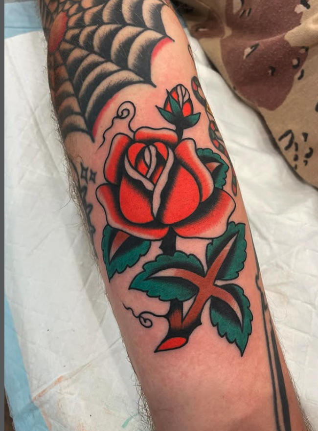 Tattoo of a traditional style red rose with green leaves and a black stem on a person's arm, with a spider web tattoo near the shoulder.