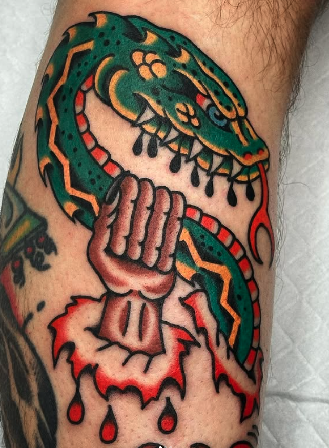 Tattoo of a green snake with yellow and red accents and fangs wrapped around a clenched fist breaking through a red and black cracked surface.