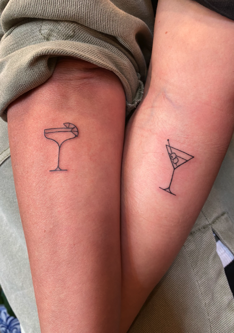 Two arms with matching minimalist line art tattoos of cocktail glasses, one with a slice of lemon and the other with an olive and lemon wedge.