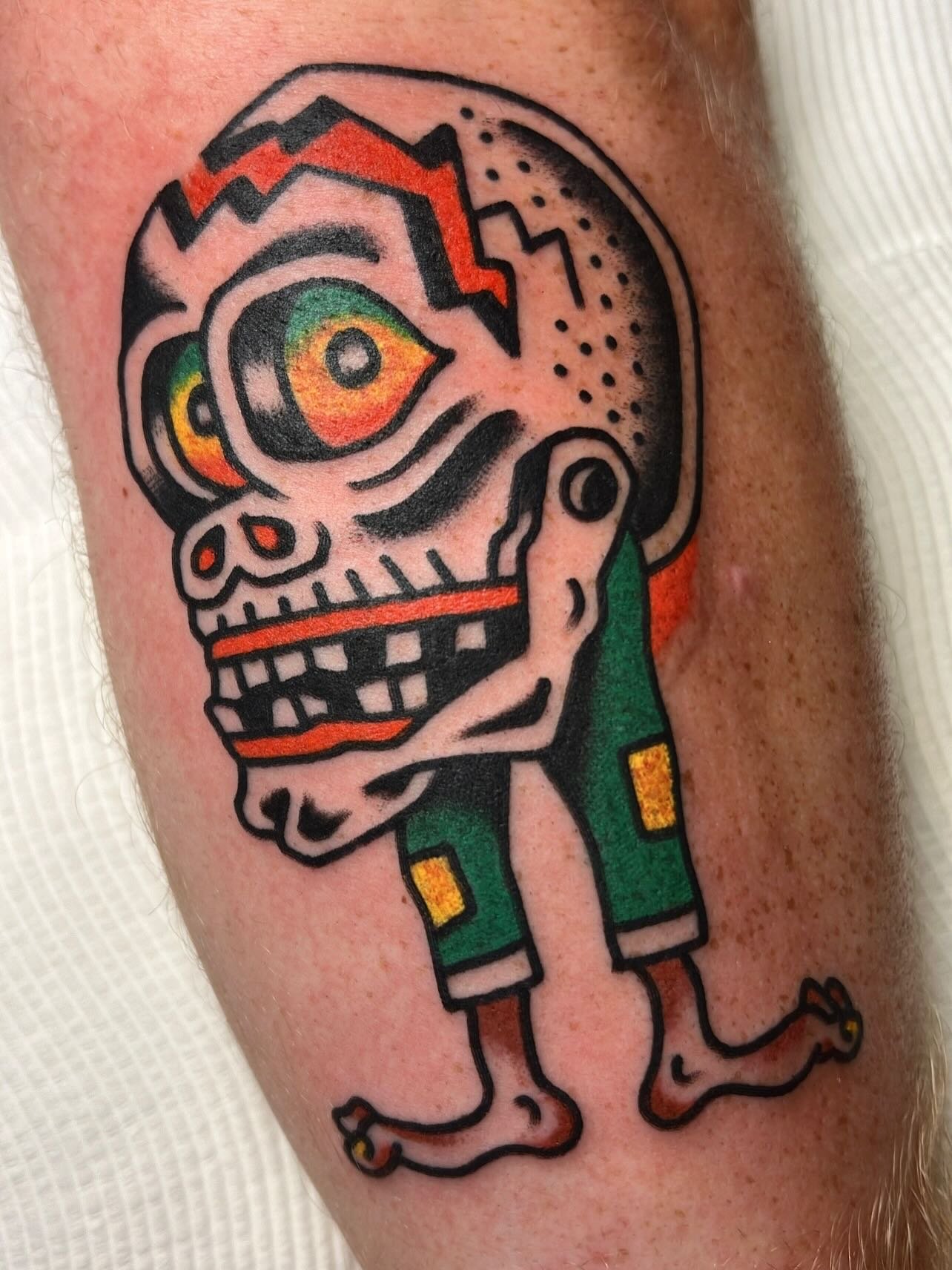 A tattoo of a colorful, cartoonish skull with large eyes, wearing green shorts, and long skinny legs with small feet, on a person's arm.