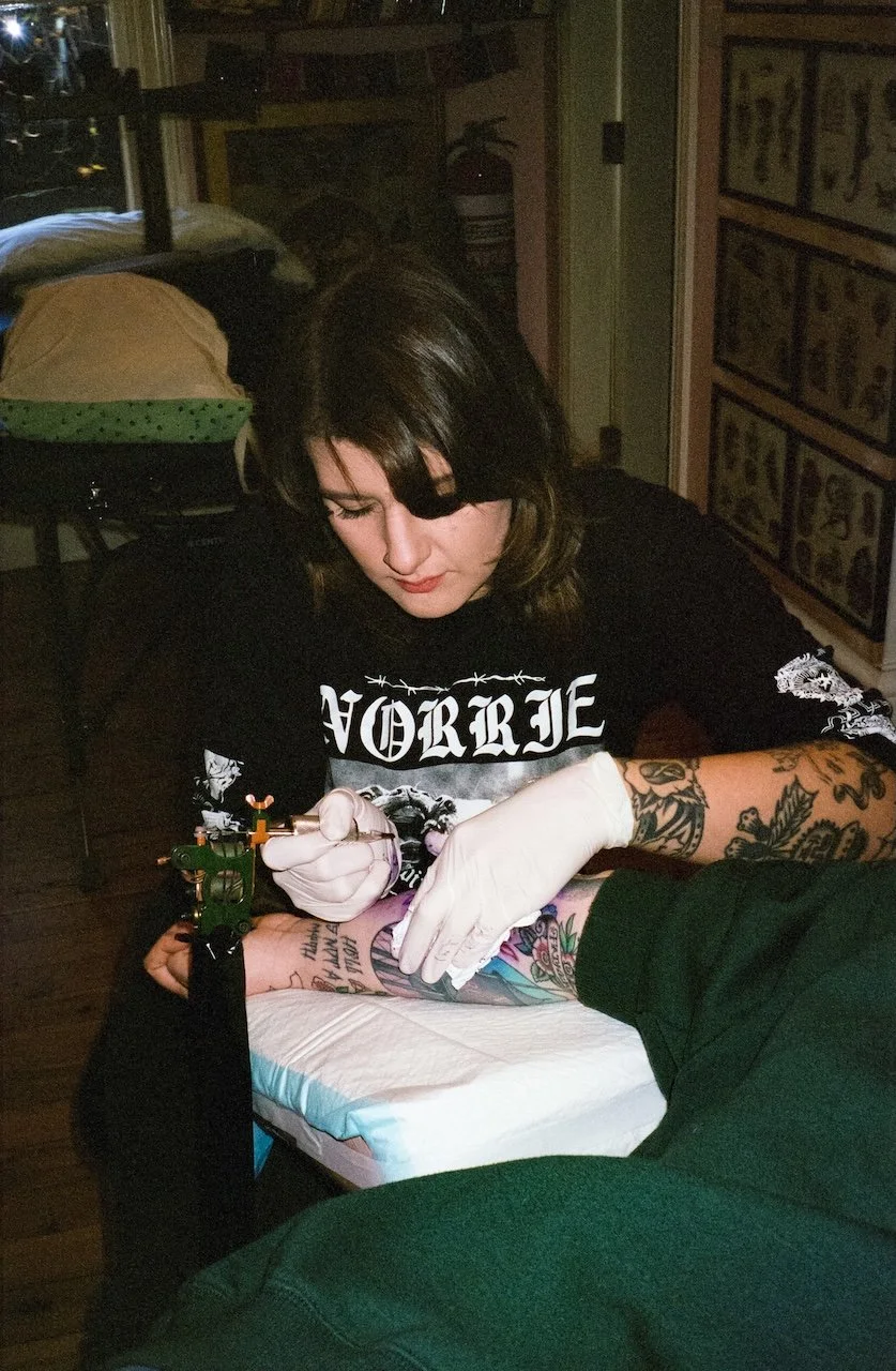 A woman with dark hair and tattoos on her arms is tattooing someone's arm in a tattoo shop. She is wearing gloves and focused on her work.