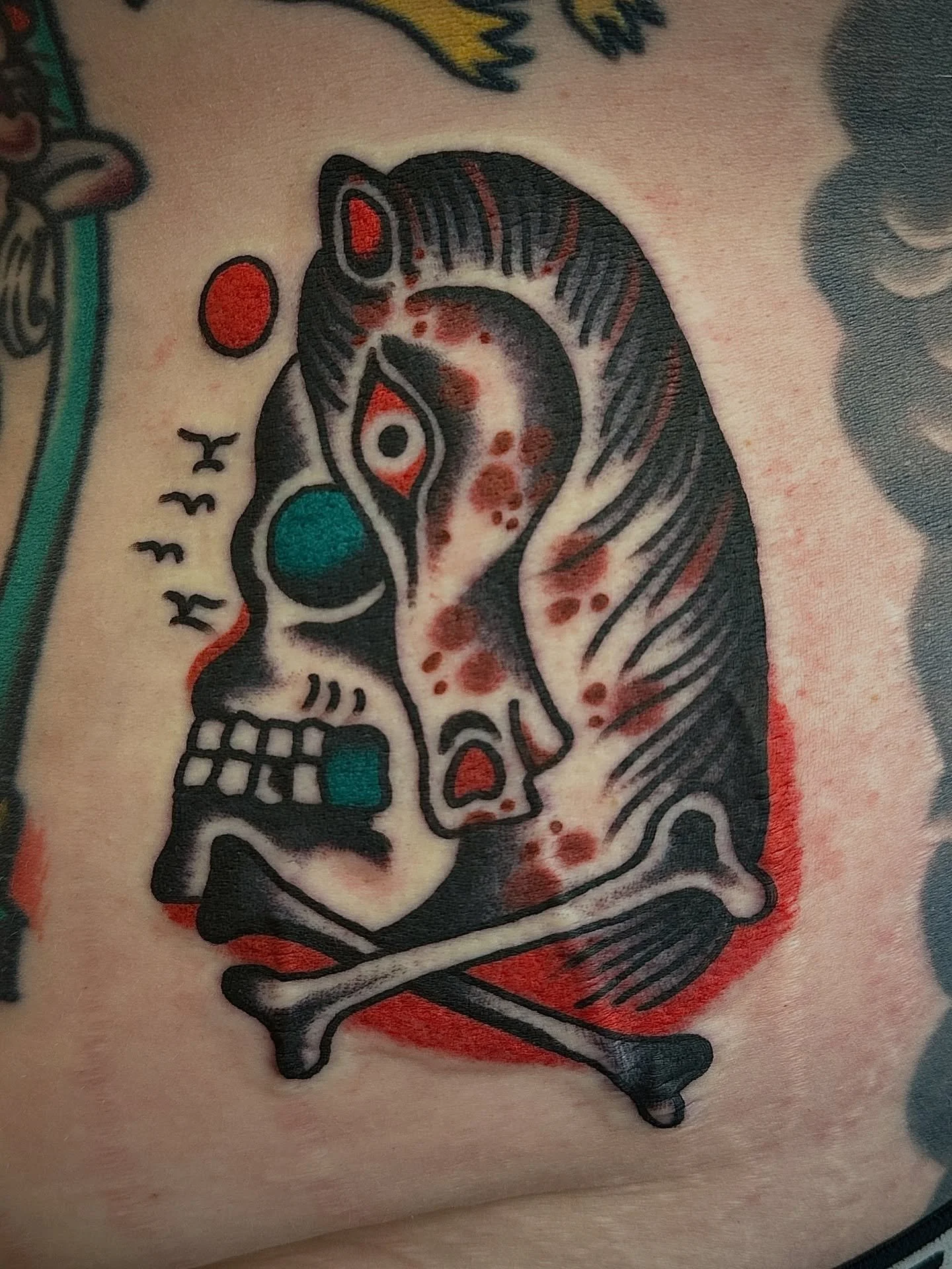 Tattoo of a stylized skull and dog skeleton with colorful elements like red, teal, and gray, surrounded by red shading.