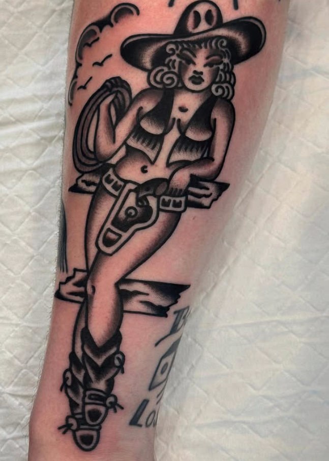 Tattoo of a woman dressed in cowboy attire, including a large hat, cowboy boots, and a belt with a buckle, standing by a fence with a scenic background of clouds and birds.