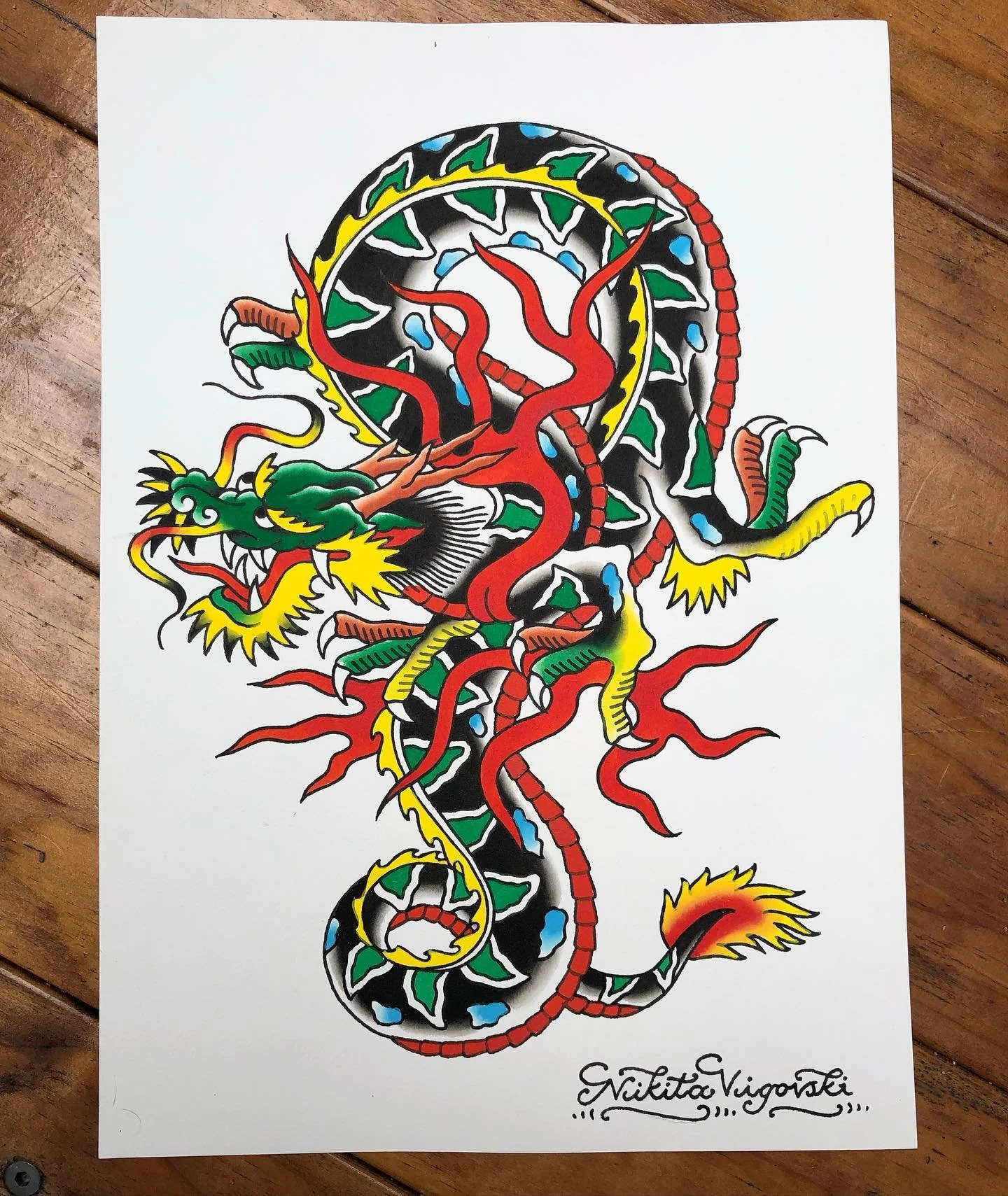 Colorful tattoo-style illustration of a dragon with a serpent body, sharp claws, and a fierce expression, signed by Nikita Vigoraski.