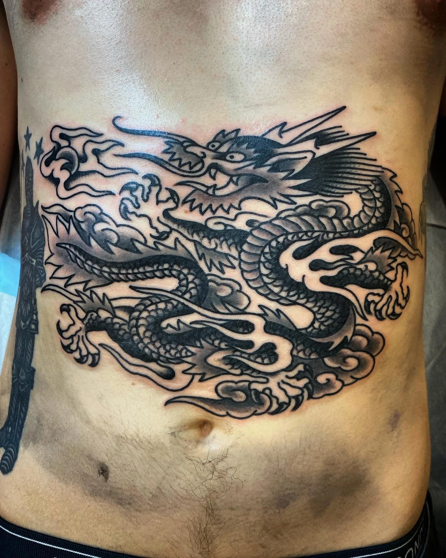 Black tattoo of a traditional Asian dragon with clouds on a person's chest.