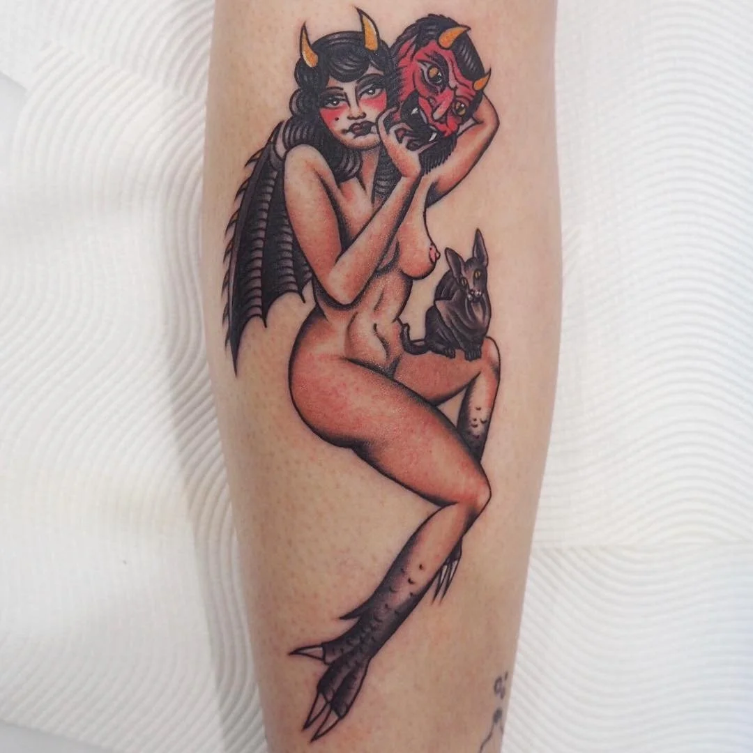 Tattoo of a mythological creature with a woman's upper body and a fish's lower body, holding a devil mask, with wings and a small black dog sitting on its lap.
