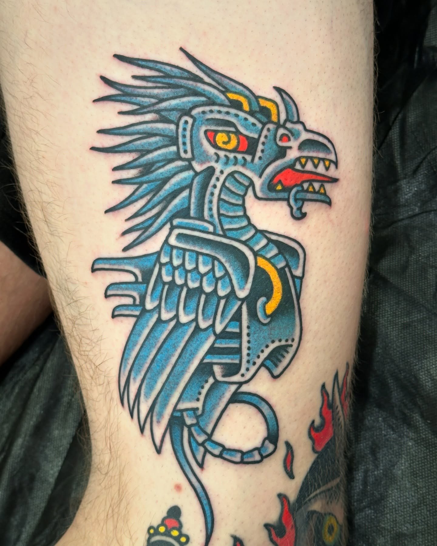 Tattoo of a mythological creature with a dragon, eagle wings, a fierce face, and flames around it.