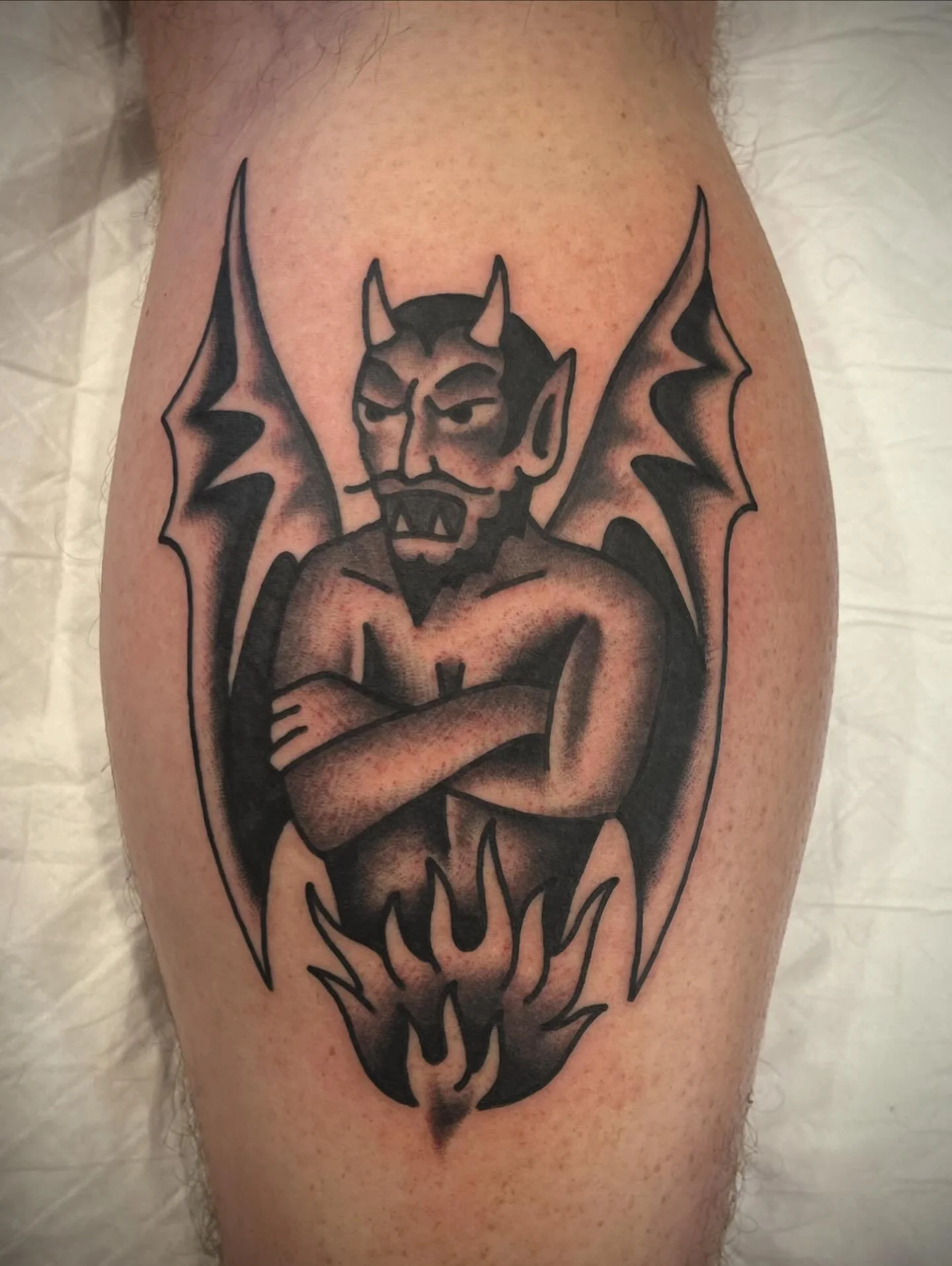 Tattoo of a devil-like creature with bat wings, crossed arms, and flames beneath, in black ink.