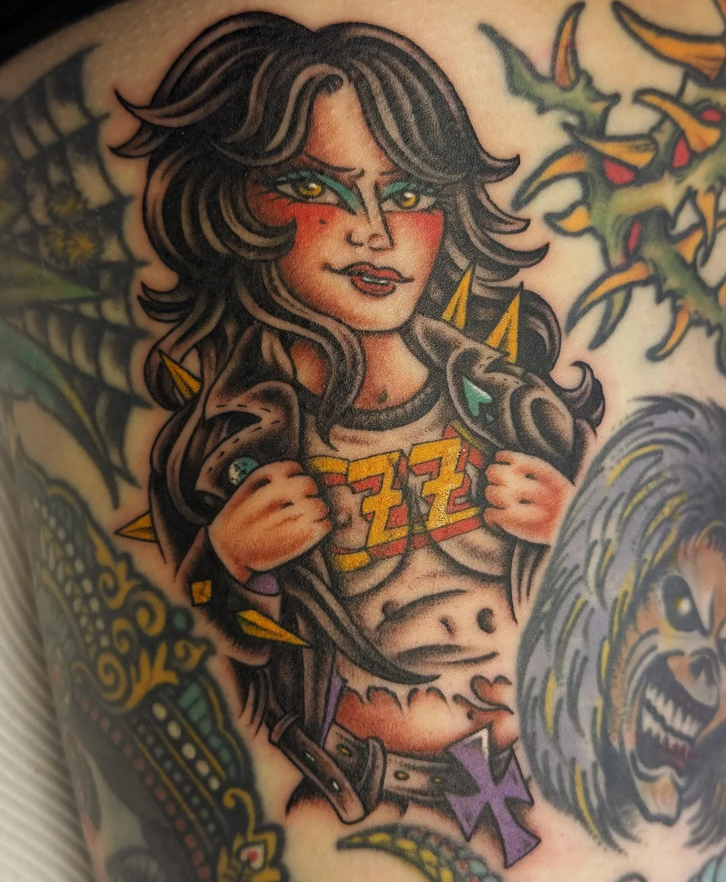 Colorful tattoo of a woman with long dark hair, holding her jacket open to reveal her midriff and a letter "Z" on her chest, with a lion's head tattoo and a spiderweb tattoo nearby.
