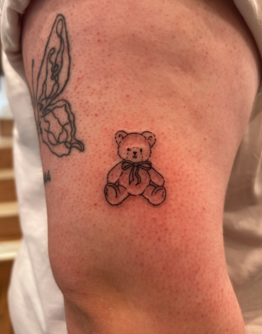 A small teddy bear tattoo with a ribbon around its neck, located on a person's upper arm, with part of an outline of a butterfly tattoo visible nearby.