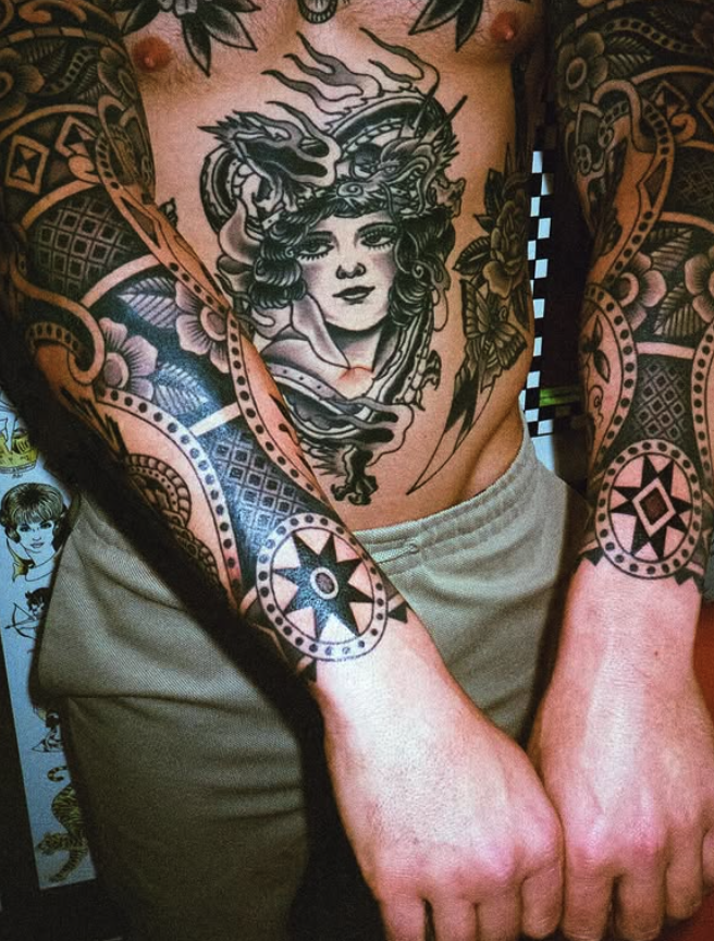 A person with extensive black tattoos on arms and chest, featuring intricate geometric patterns, a woman with flowing hair, a dragon, and floral designs.