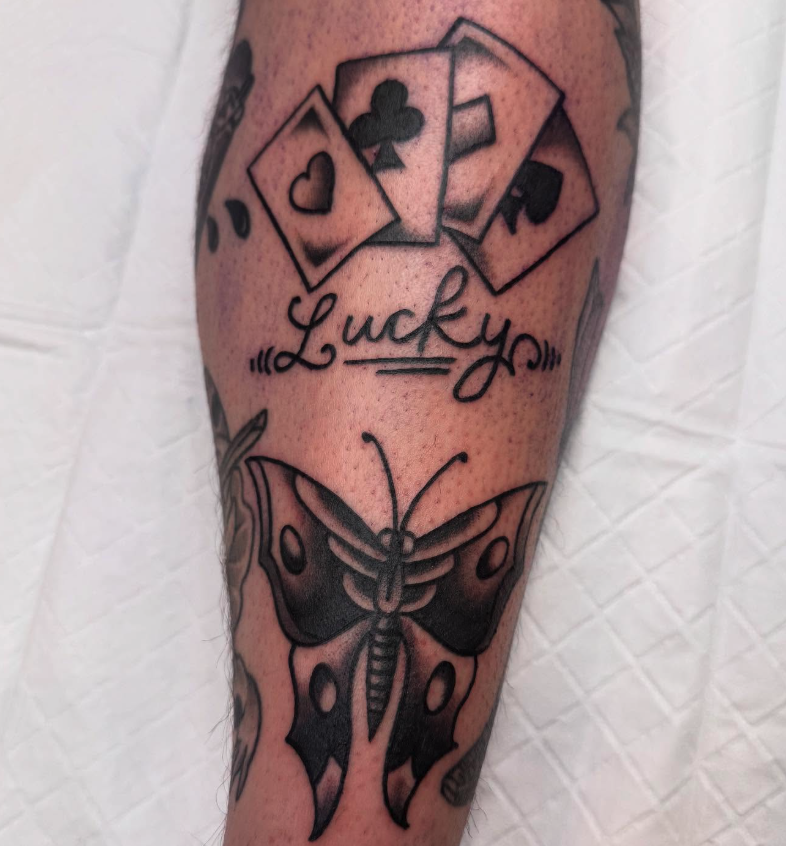 Tattoo of four playing cards, the word "Lucky" with decorative scrolls, and a butterfly with black and white wings on a person's arm.