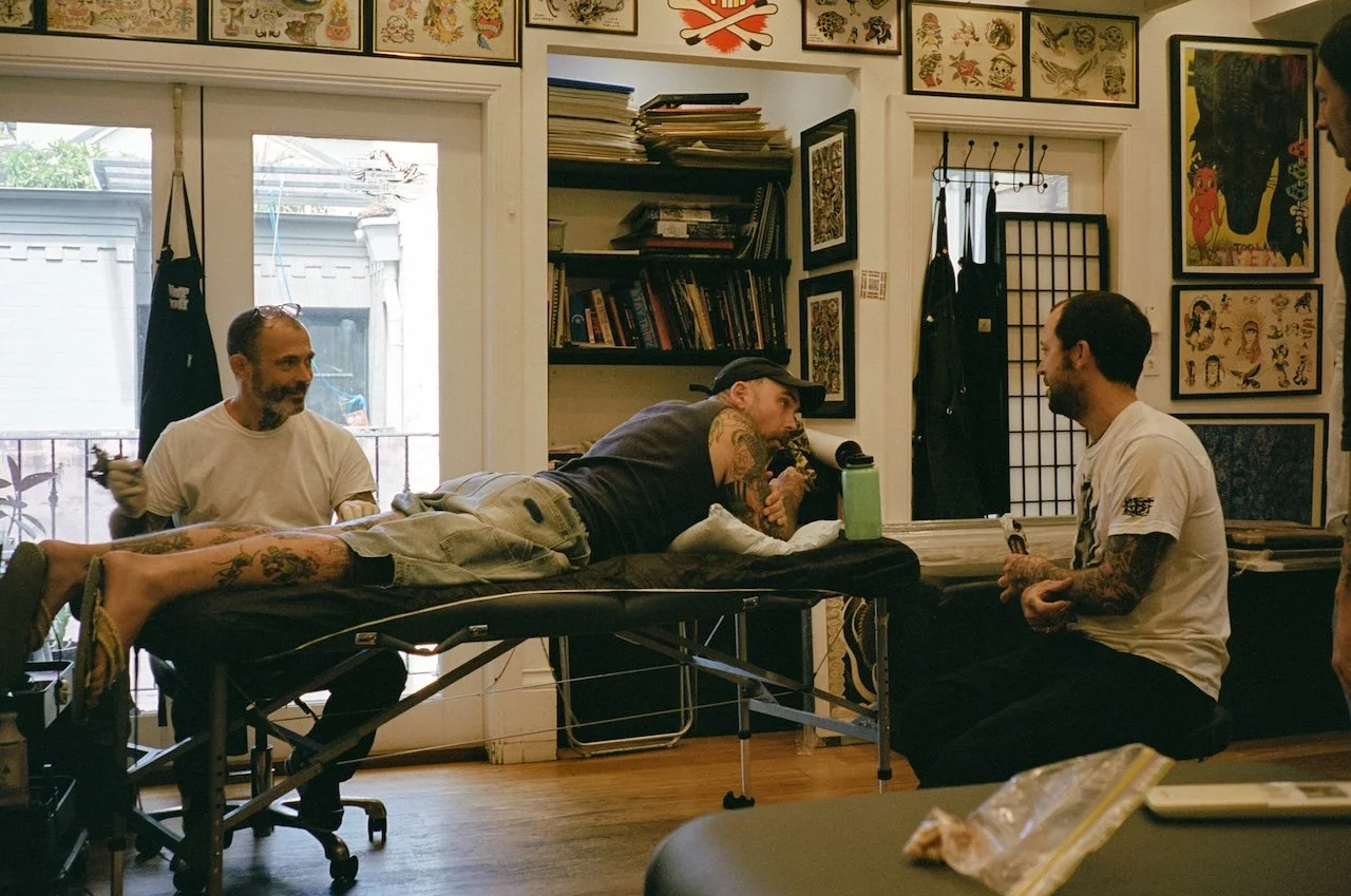 A tattoo artist giving a tattoo to a client lying on a tattoo table in a tattoo shop, with two other men nearby talking and smiling, surrounded by tattoo art on the walls.