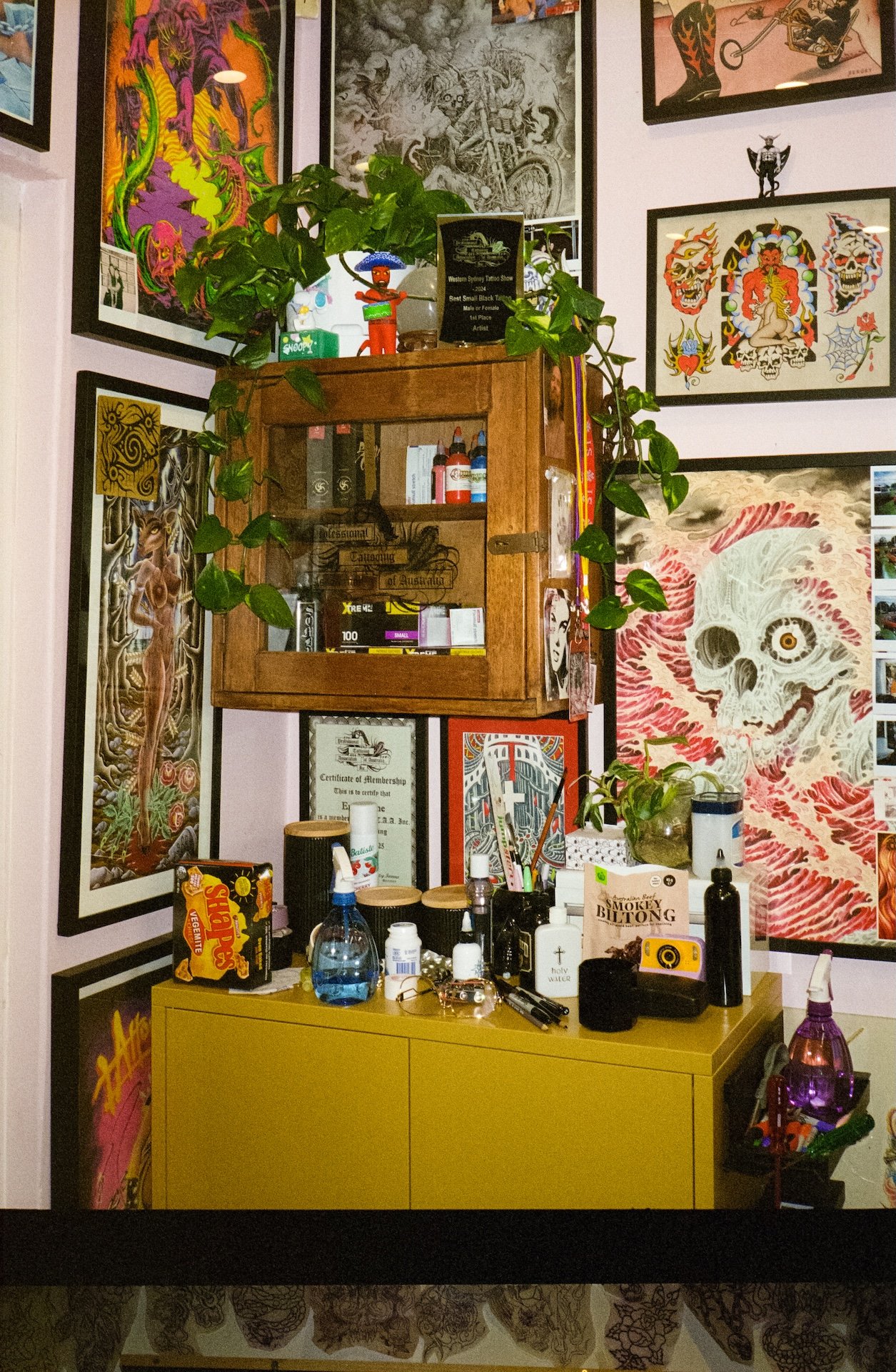 A colorful room with various framed artwork on the walls, a yellow cabinet, and a small wooden cabinet with a glass door. Items on the yellow cabinet include bottles, a spray bottle, a box of Side Trams, and personal items. The artwork on the walls f