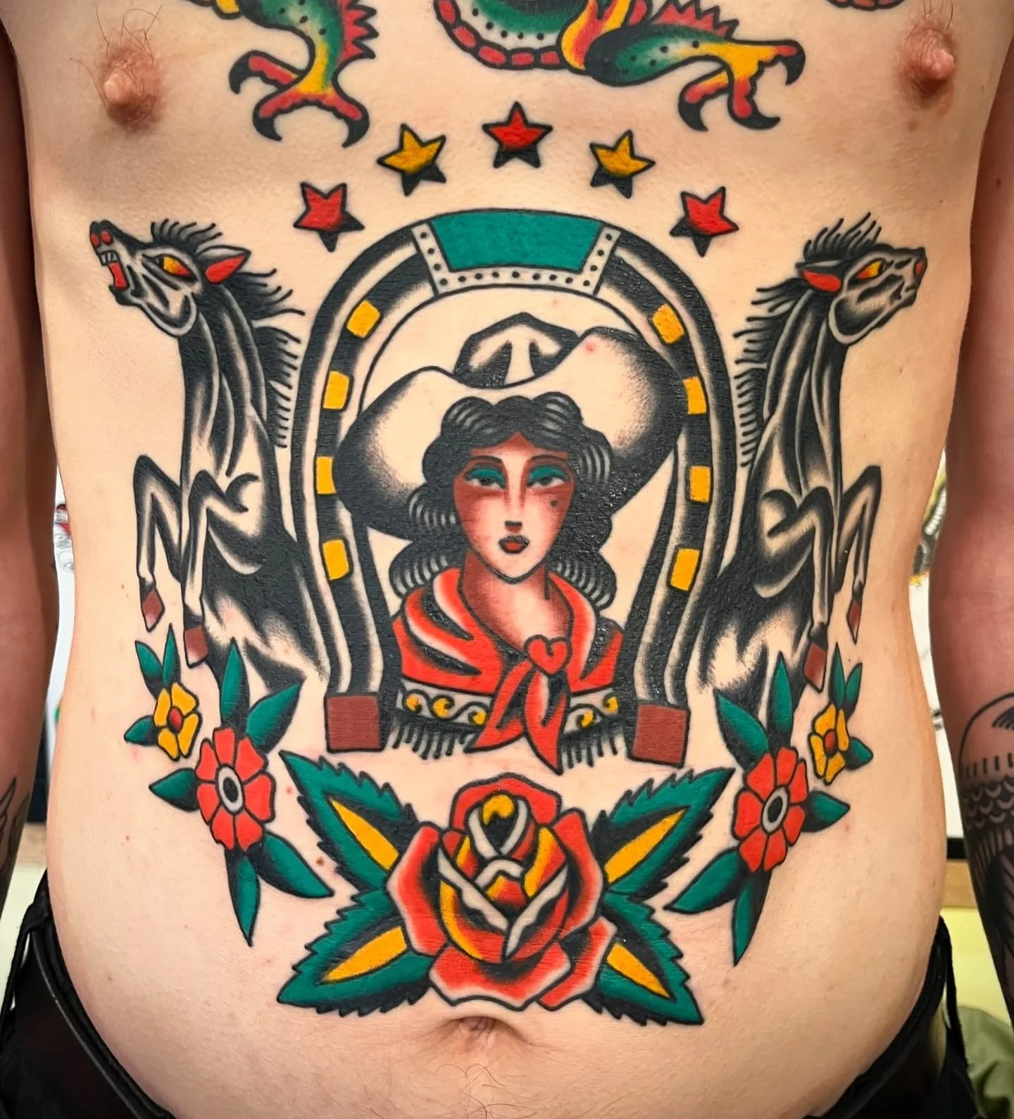 Colorful tattoo on a person's chest featuring a woman with dark wavy hair, a large hat, and a red scarf, surrounded by two black and white horses, flowers, stars, and a rose at the bottom.