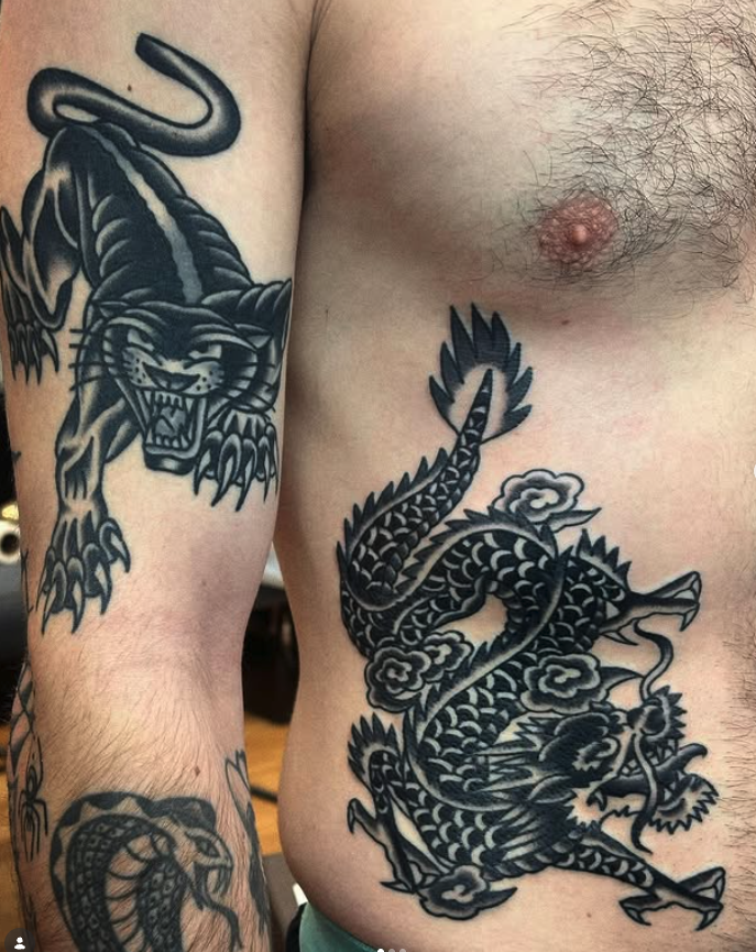 Close-up of a person's chest and shoulder with multiple black ink tattoos, including a panther on the shoulder and a dragon on the side of the torso.