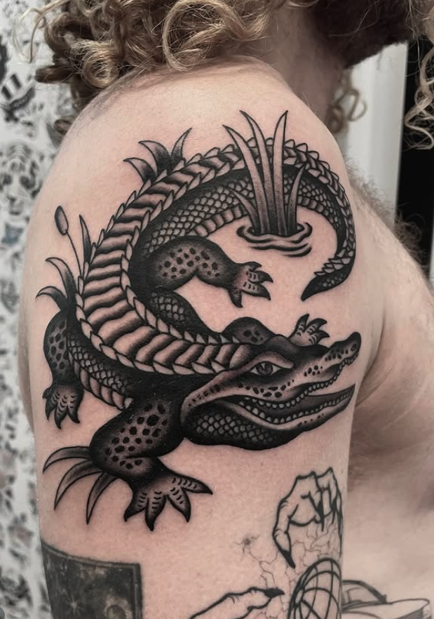Tattoo of a crocodile and a snake intertwined on a person's shoulder, in black ink.