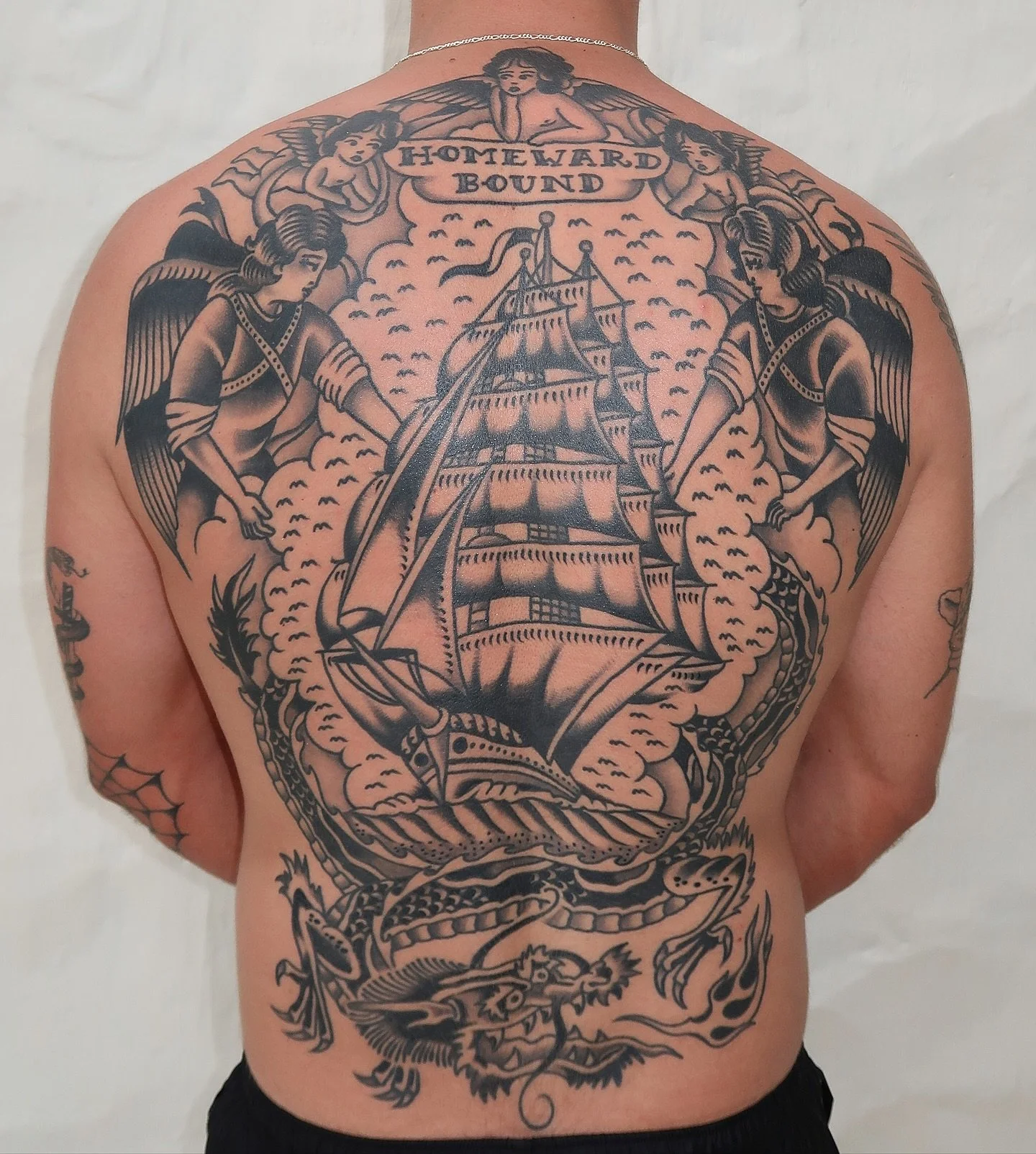 Tattoo on person's back showing a sailing ship among clouds, with angels or cherubs and a dragon at the bottom, with the words "Homeward Bound" at the top.