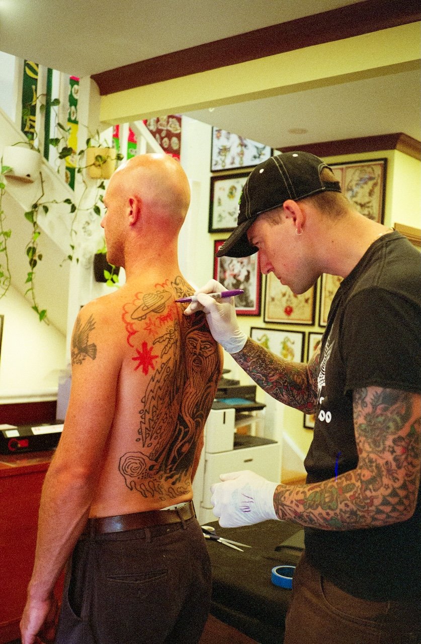 A tattoo artist with a sleeve full of colorful tattoos is tattooing a large, detailed design on a man's back in a tattoo studio. The man has a shaved head and is shirtless, wearing dark pants and a belt. The studio decor includes framed tattoo artwor