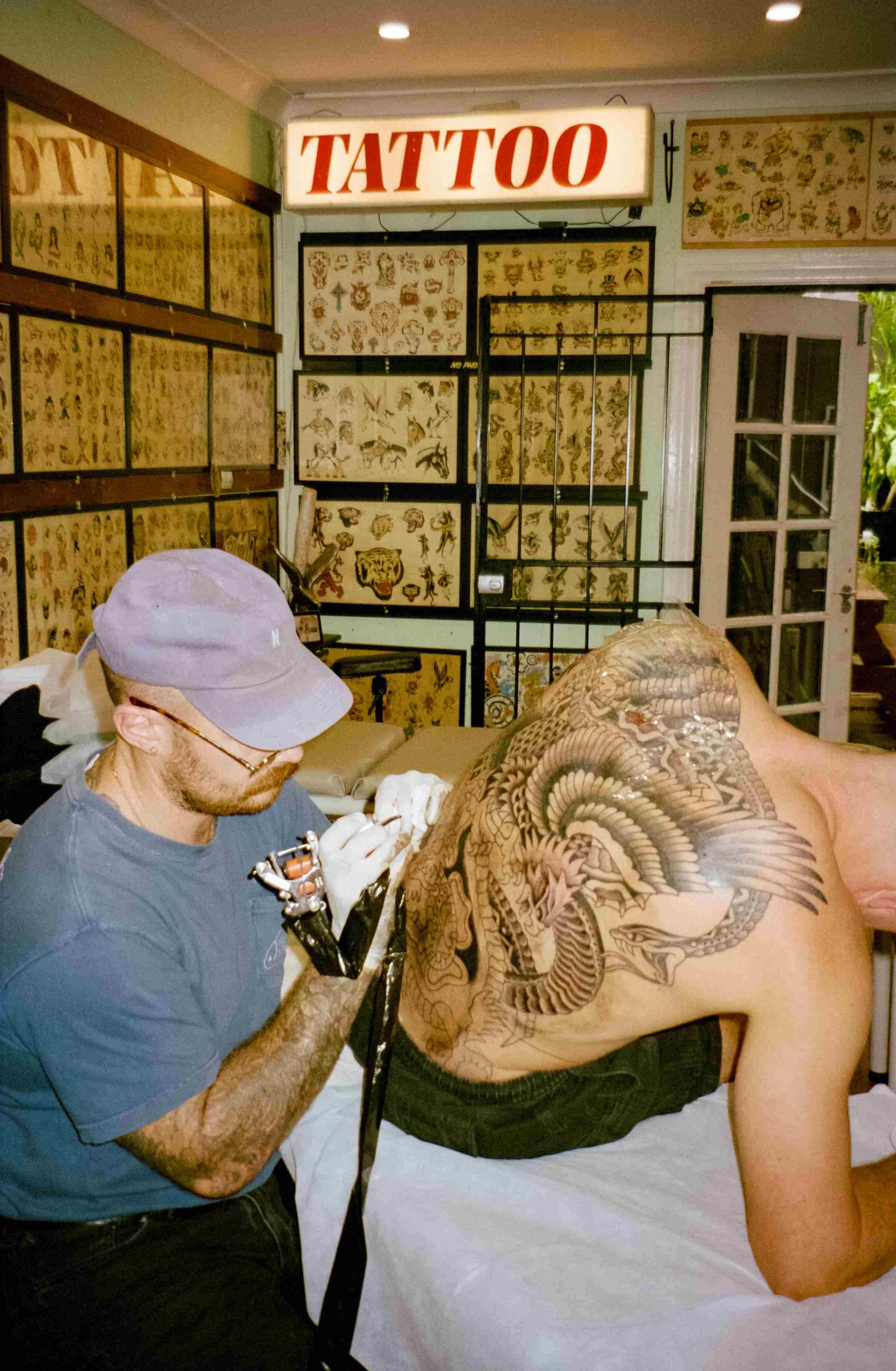 A tattoo artist is working on a large intricate dragon tattoo on a man's back in a tattoo shop, with tattoo flash sheets on the walls and a lit 'TATTOO' sign above.