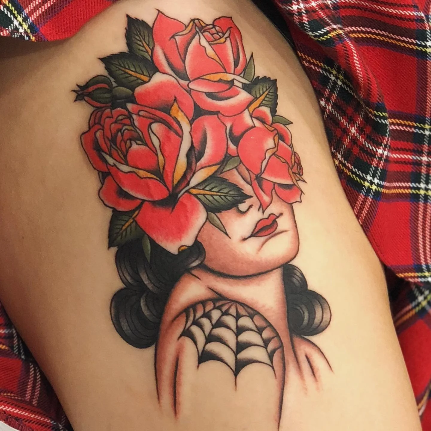 A tattoo of a woman's face with her eyes closed, surrounded by pink roses and green leaves on her head. She has dark, curly hair and red lips. A spiderweb design is on her shoulder.