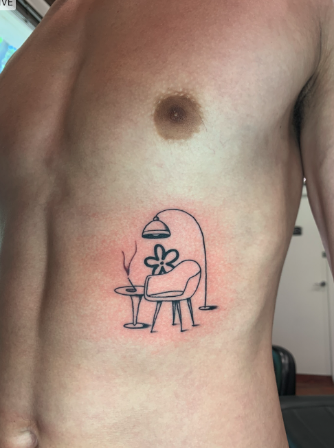 Tattoo of a simple line drawing of a chair with a flower as a backrest, a floor lamp, and a small table with a cup and smoke, located on a person's upper chest.