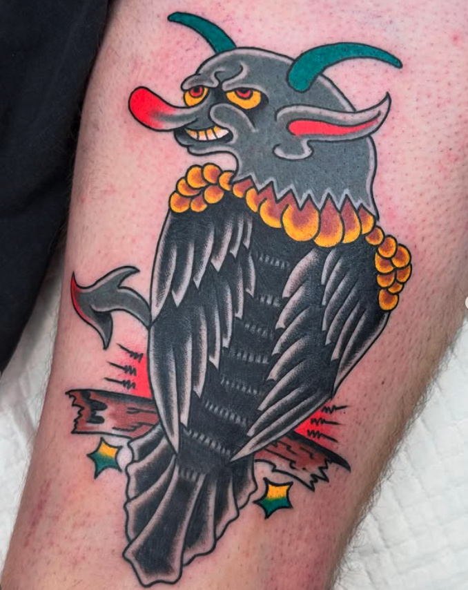 Traditional-style tattoo of a grumpy goat with blue horns, red nose, and a beaded necklace, emerging from a cracked, dark-colored rock with colorful stars surrounding it.