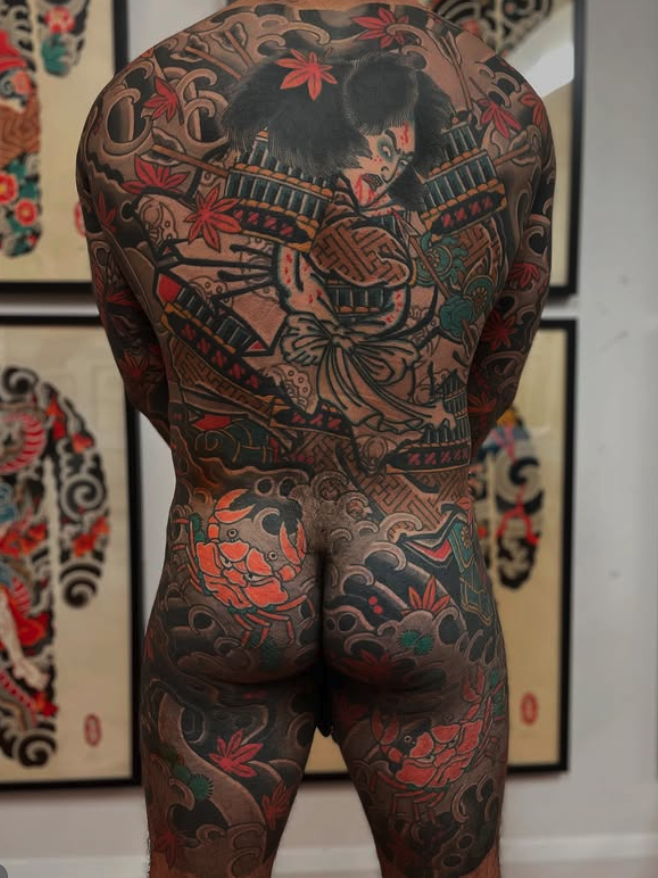 Back view of a heavily tattooed person with traditional Japanese-style tattoos on their back and legs, displaying various colorful themes including a samurai, clouds, and floral patterns.