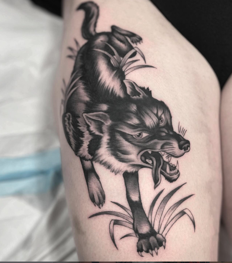 Black and gray tattoo of a fierce wolf with glowing eyes, bared teeth, and a snarling expression, surrounded by grass.