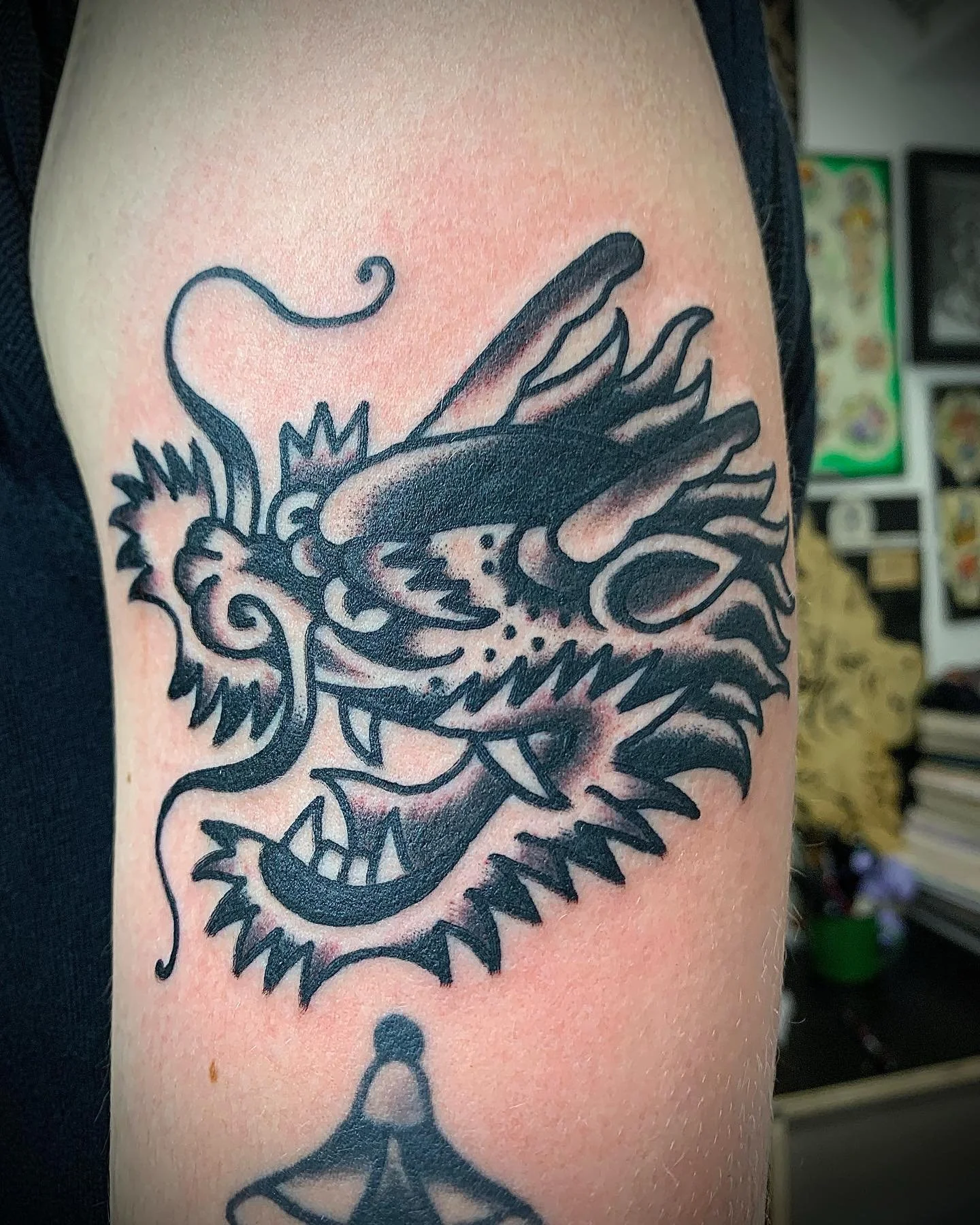 Black tattoo of a stylized dragon's head with sharp teeth, flames, and a curled tongue on person's upper arm.