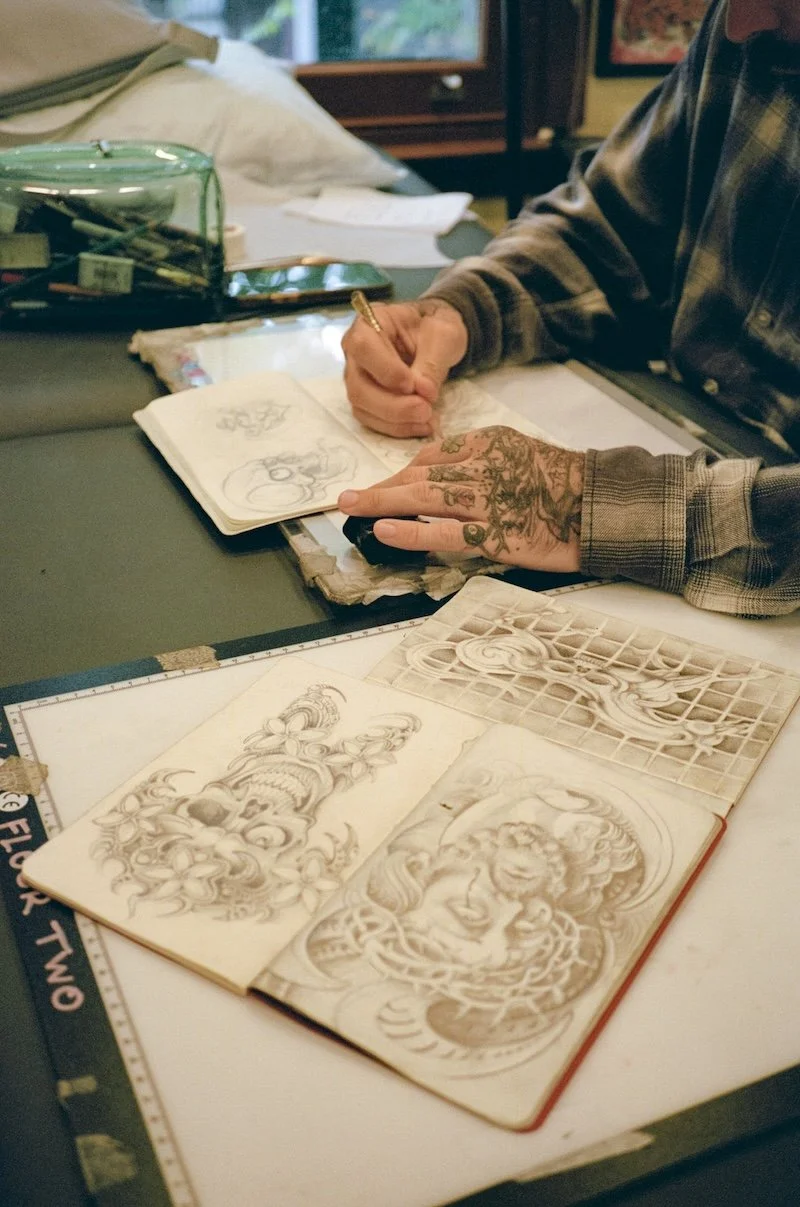 Person drawing at a desk surrounded by tattoo sketches and tattoo flash sheets.