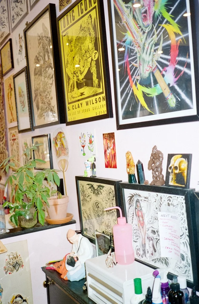Gallery wall with colorful artwork and posters, including a bright yellow poster for S. Clay Wilson, a vivid anime-style poster, and various smaller art pieces. A table with plants, figurines, a pink bottle with a straw, and art supplies is in front 