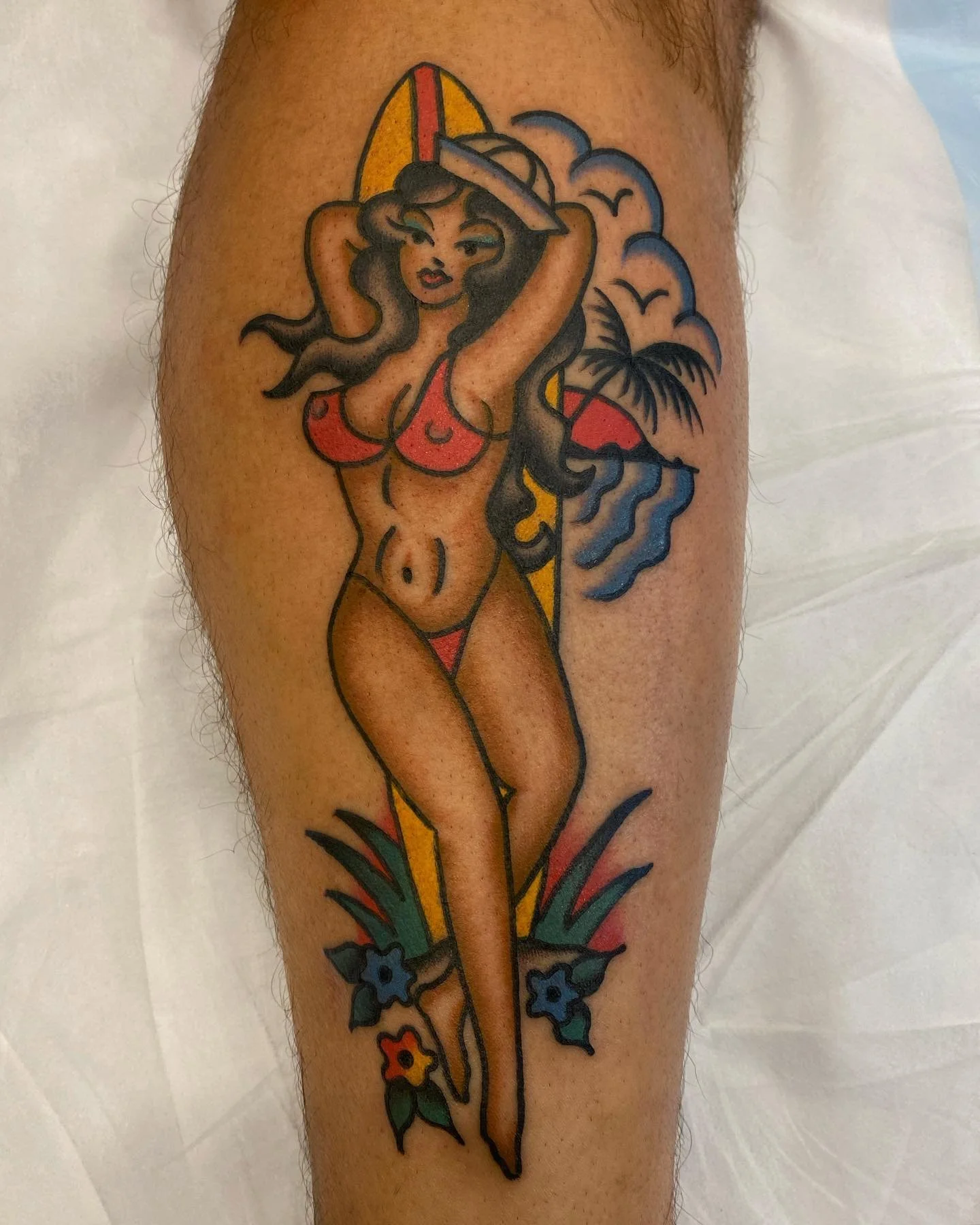 Traditional tattoo of a woman in a red bikini holding a hat, standing in front of a surfboard, with palm trees, clouds, birds, waves, and flowers in the background.