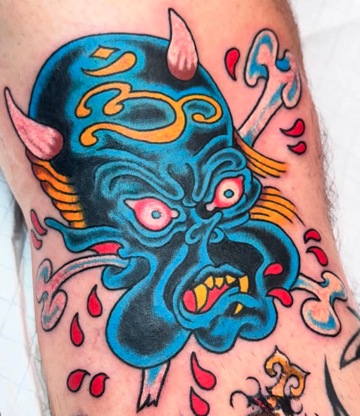 Colorful tattoo of a blue demon with pink eyes, yellow teeth, sharp horns, and flowing hair, surrounded by drops of red ink.
