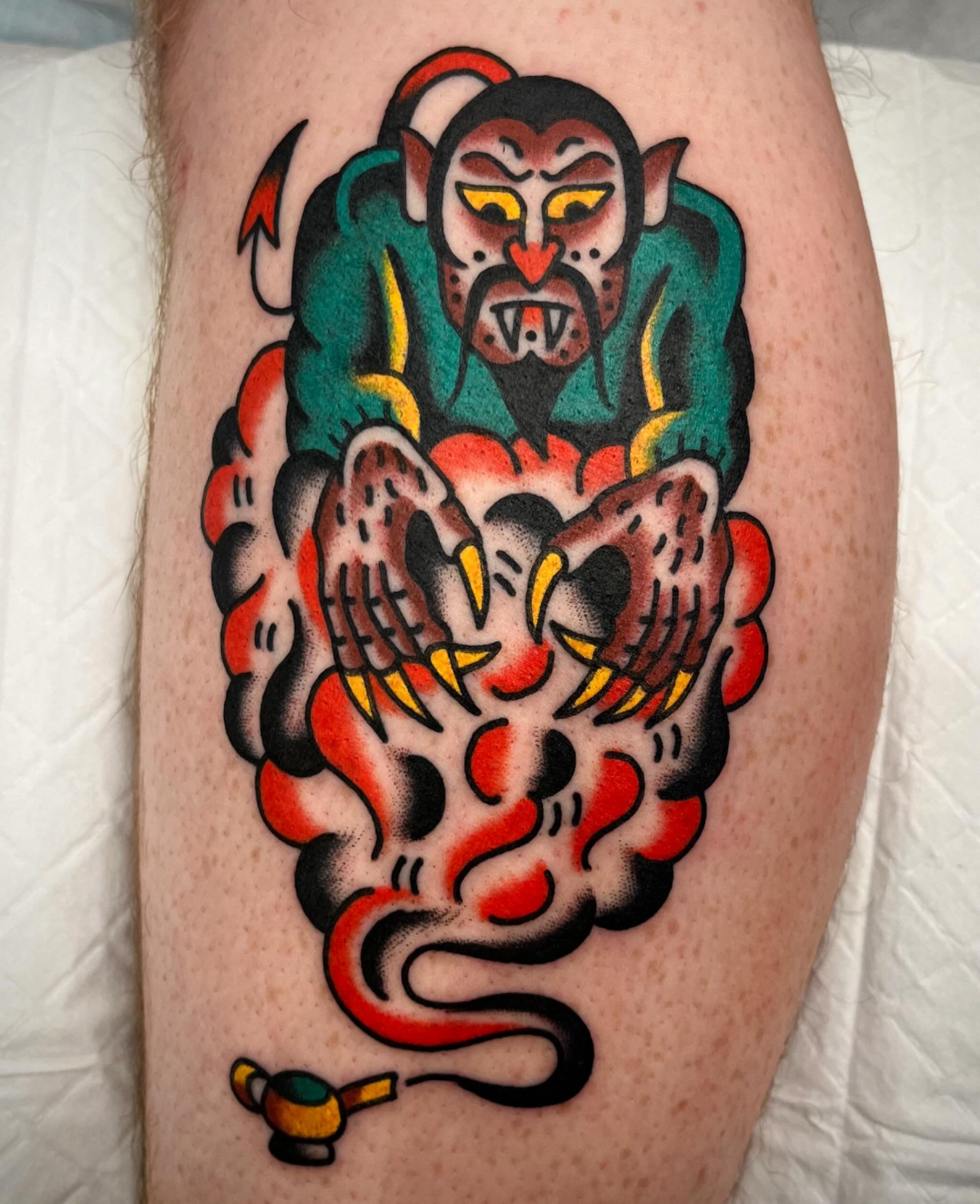 Tattoo of a demon with a fierce expression, sharp teeth, and pointed ears, holding a grey object with a yellow handle, with a cloud of red and black smoke beneath.