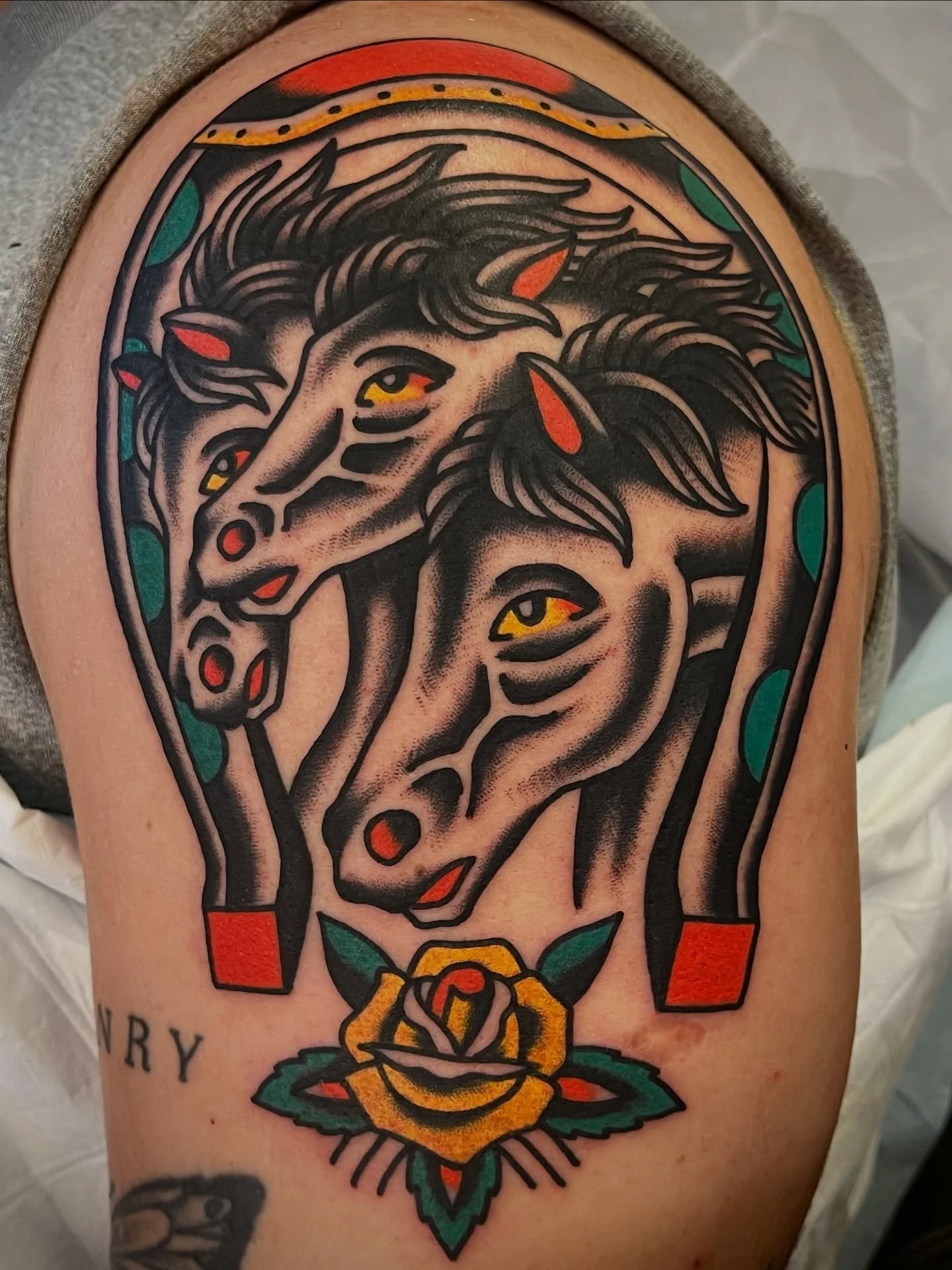 Colorful tattoo of two horses' heads with yellow eyes, surrounded by teal, orange, and red accents, and a yellow rose at the bottom.