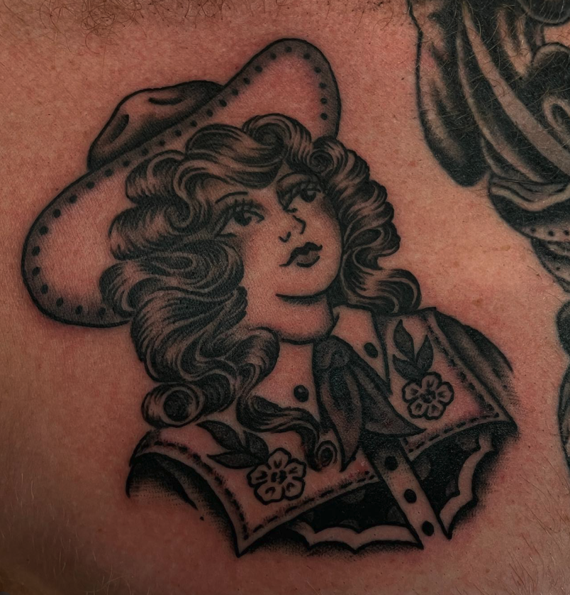 Tattoo of a woman with curly hair wearing a wide-brimmed hat, a vest with floral embroidery, in a traditional black and gray style.