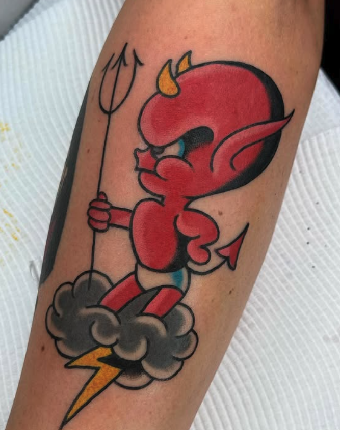 A colorful tattoo of a cartoon devil holding a trident, standing on a cloud with a lightning bolt beneath. The devil is red with yellow horns, blue eyes, and a pointed tail.