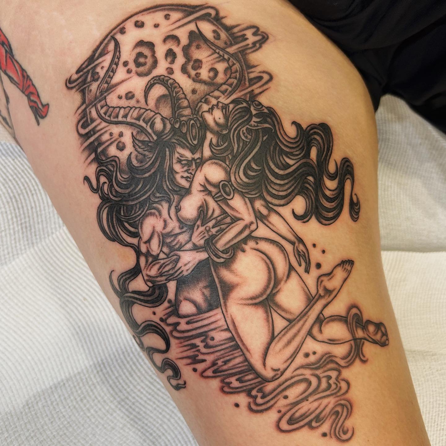 A black and gray tattoo of a mythological scene featuring a woman with long flowing hair, wearing jewelry and a revealing outfit, kneeling with one leg in the water, holding a spear, with a creature resembling a demon or demon-like figure behind her.