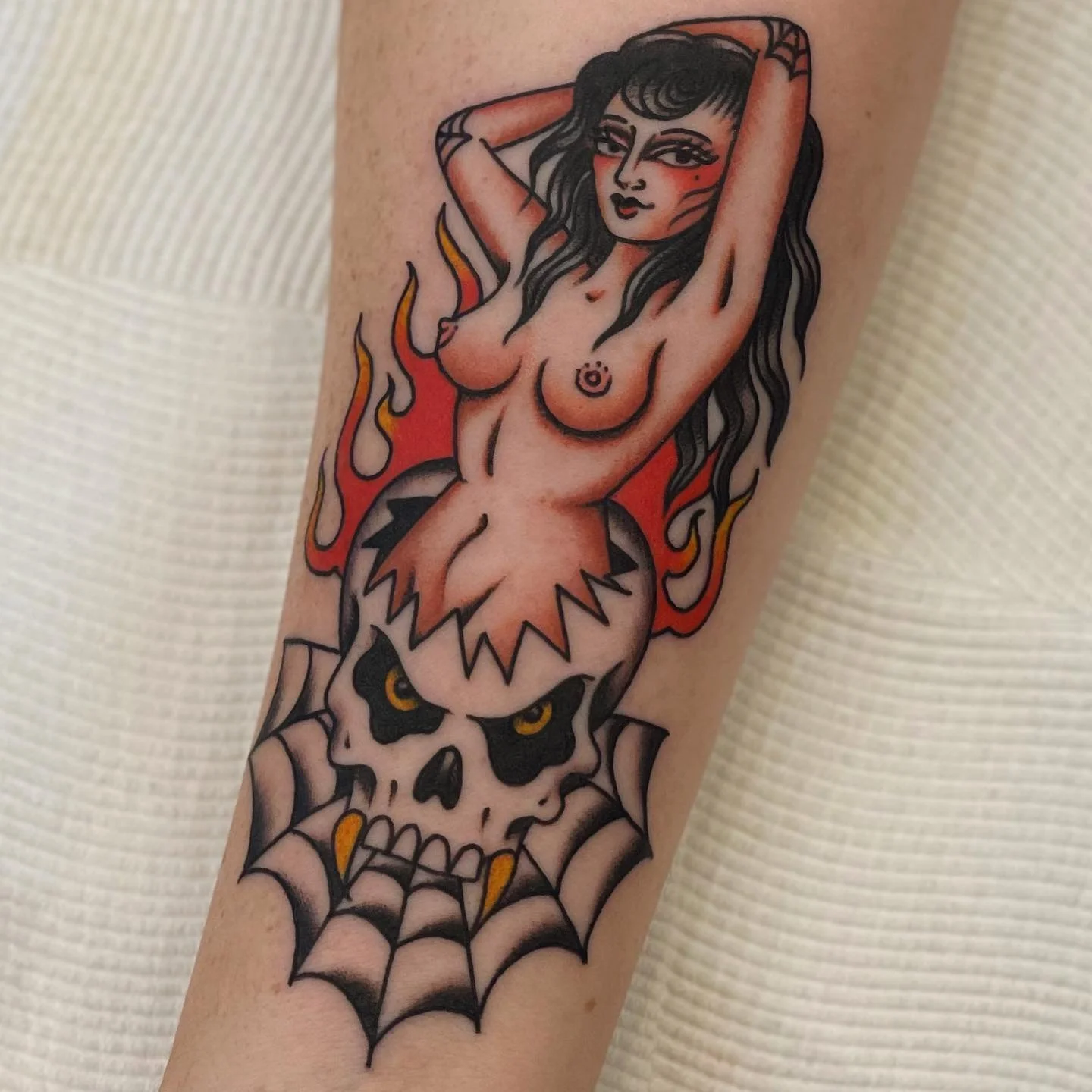 Tattoo of a woman with long black hair, posing with arms above her head, above a skull with yellow eyes, surrounded by flames and a spider web at the bottom.