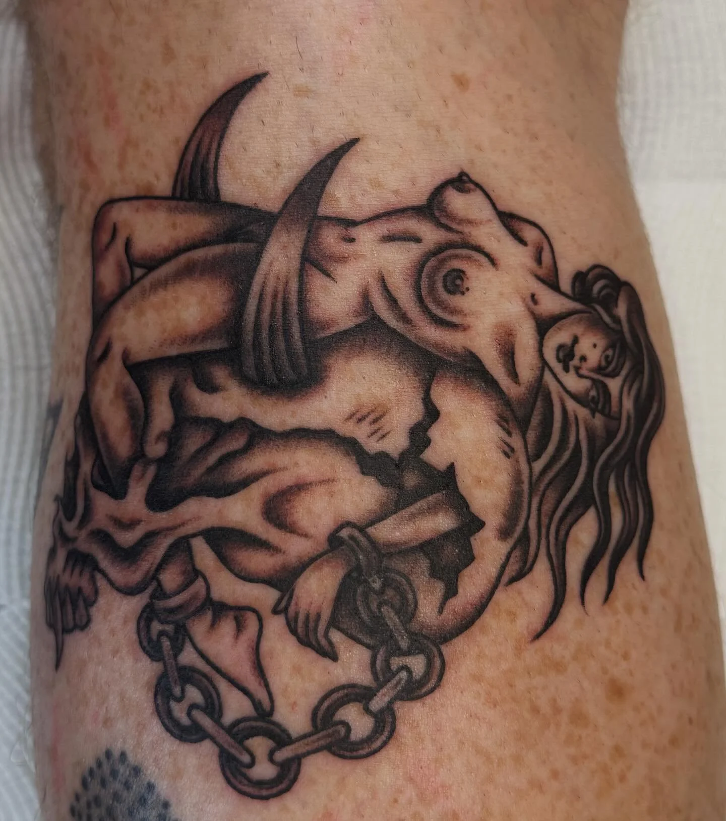 Tattoo of a woman with long hair, chained and kneeling, with a mixture of realistic and stylized art in black and gray ink.