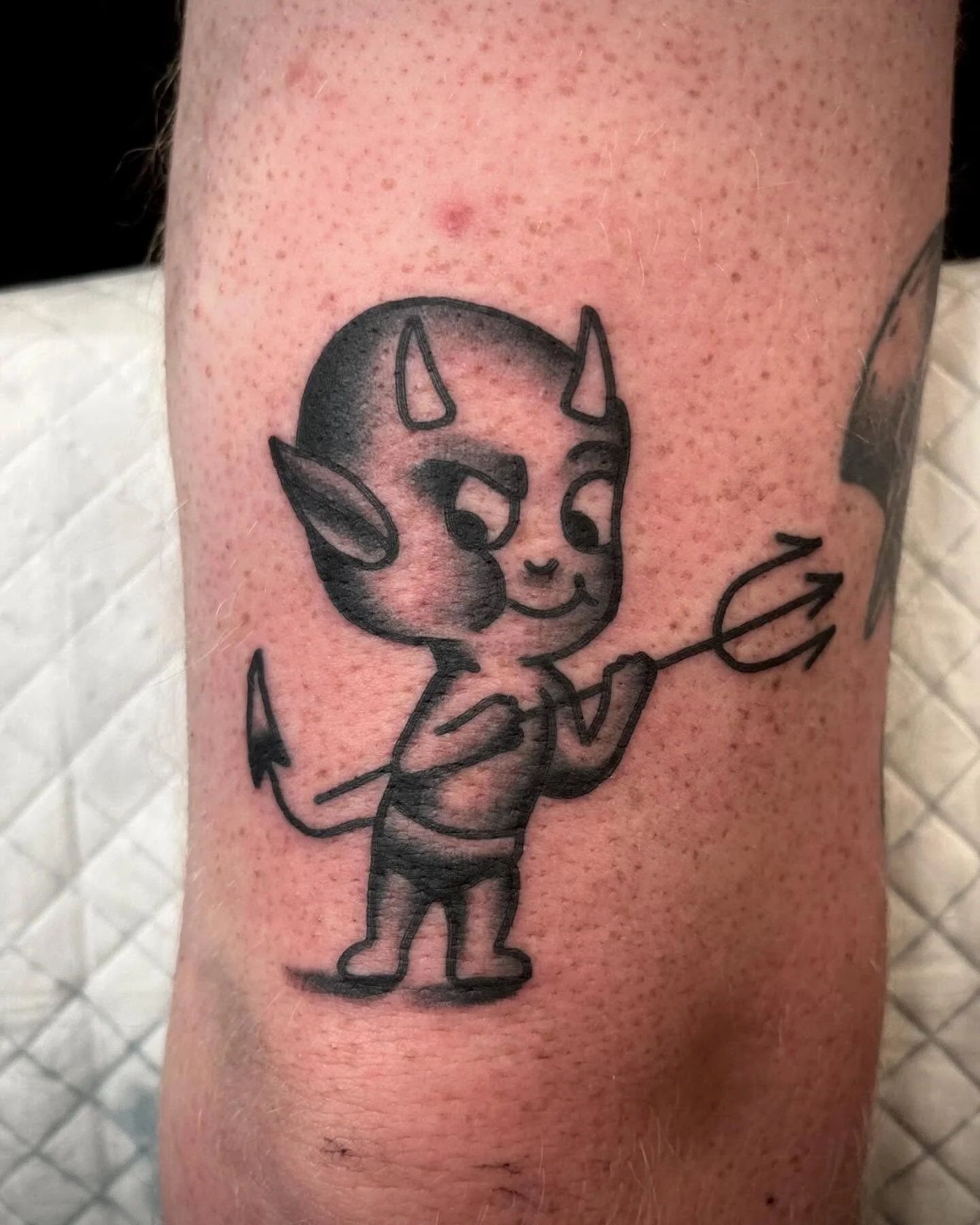 Tattoo of a cute, cartoon-style devil with horns, a tail, and a small spear, standing with a happy expression.