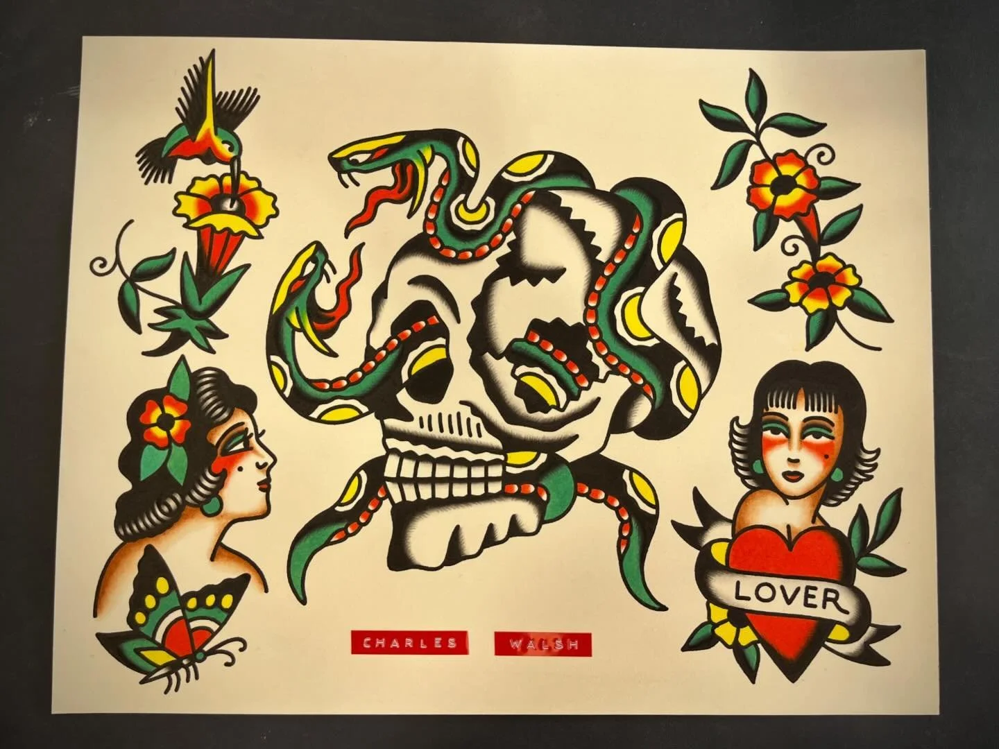 Traditional tattoo featuring a skull with a snake wrapped around it, surrounded by colorful flowers, a butterfly, a woman with a flower in her hair, and a woman with a heart labeled "LOVER".