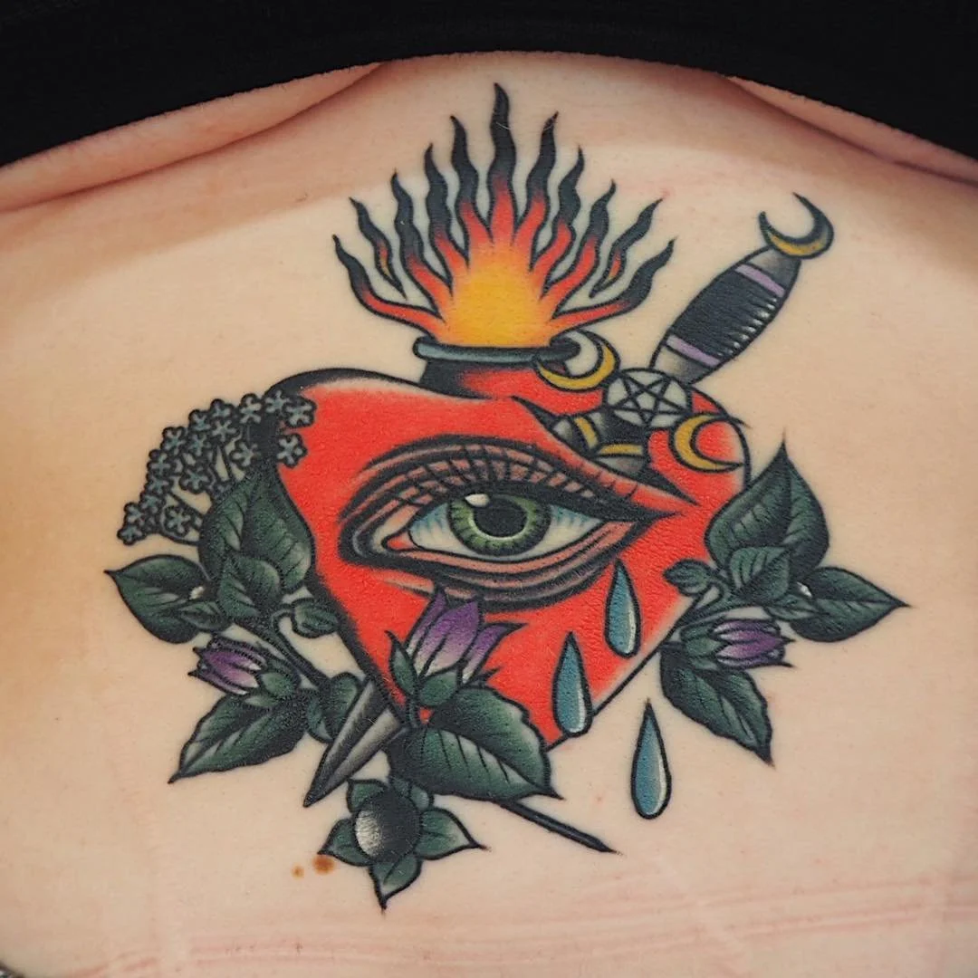 Tattoo of a heart with a single eye, a dagger piercing through, surrounded by leaves and purple flowers, with a flame and a knife above.
